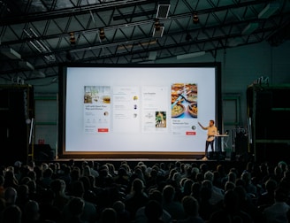 person discussing while standing in front of a large screen in front of people inside dim-lighted room
